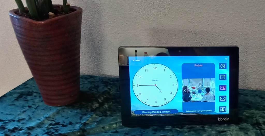 Je ziet een kast, met een groen kleedje erop. Aan de linker kant staat een plant in een hoge pot. Daarnaast staat de BBrain, een scherm. Daarop zie je een klok die de tijd aangeeft: het is kwart voor 5, en een foto van twee vrouwen die bij een tafel met spullen staan. Achter de tafel op de foto staat een andere vrouw uitleg te geven over de spullen.