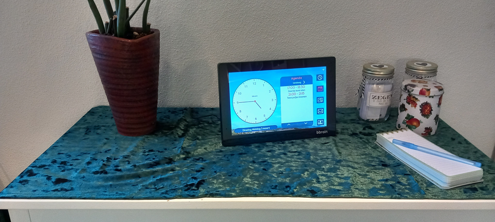 Je ziet een witte kast met daarop een groen kleedje. Op het midden van de kast staat een BBrain dementieklok. Aan de rechterkant zie je een aantal potjes en een schrijfblok.

Op het scherm van de BBrain zie je een klok die aangeeft dat het kwart voor 5 is. Naast de klok staan de twee eerstvolgende afspraken in beeld: Doortje komt eten om 5 uur en om 21 uur moeten twee pufjes worden ingenomen.